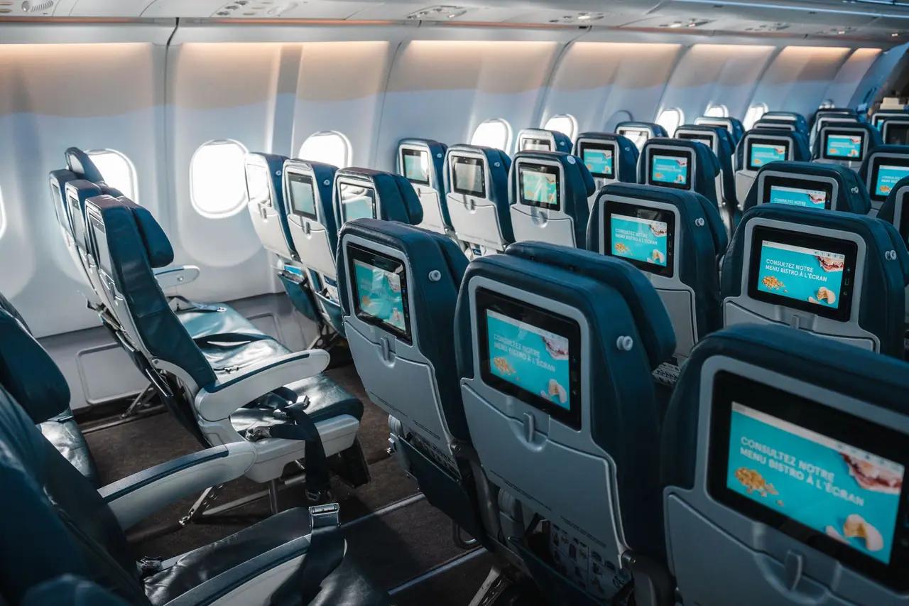 Rows of empty economy seats with seatback screens inside an airliner cabin, window shades up